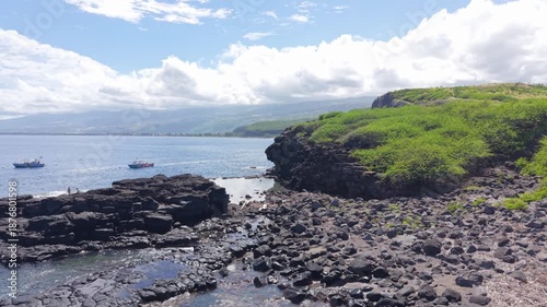 Aerial drone footage flying along the coastline and natural pools before moving toward two diving boats, passing above a snorkeler visible in the water near Reunion Island. Ideal for marine activities