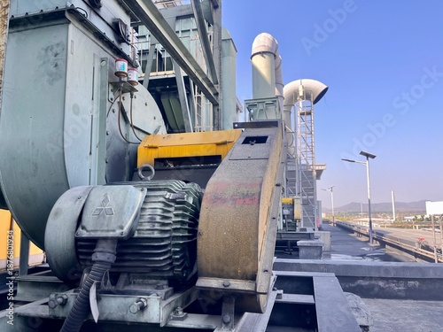 Foto Industrial centrifugal blower with electric motor and exhaust duct system installed on factory rooftop, showing ventilation equipment for air pollution control in manufacturing plant