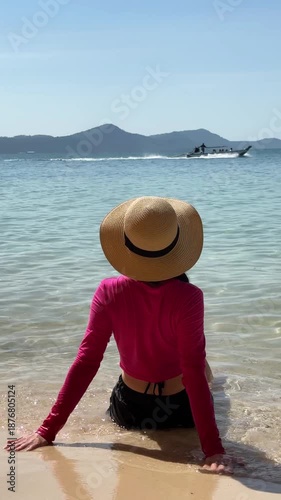 Young woman in bright clothes and straw hat sitting on the beach by the azure sea, enjoying the stunning view and gentle waves.	
