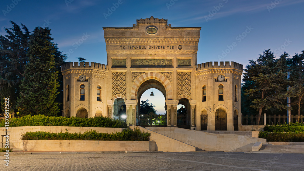 Fototapeta premium Istanbul University's historic main gate, illuminated at dusk, with ornate architecture and clocks. Istanbul, Turkey.