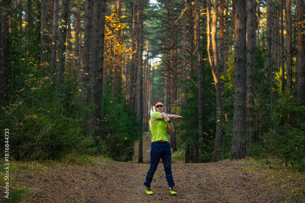 Fototapeta premium Man in athletic gear and sunglasses stretching arms on a forest trail, warming up for a run amid pine trees and autumn light, focused on fitness and well-being
