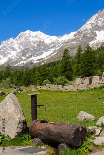 Foto Traditional wooden water fountain in an alpine meadow with green grass, stone walls, forest and snowcapped Mont Blanc mountains near Courmayeur on a sunny day