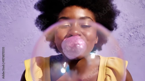 A playful young woman with an afro blows a large bubblegum bubble. The pink bubble pops messily on her face and she laughs. Fun and carefree moment against a vibrant purple wall.