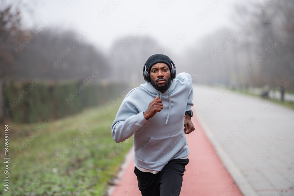 Fototapeta premium Focused Black man running outdoors wearing headphones and a hoodie on an overcast day, expressing determination, energy, and motivation, capturing fitness, endurance, and healthy active lifestyle