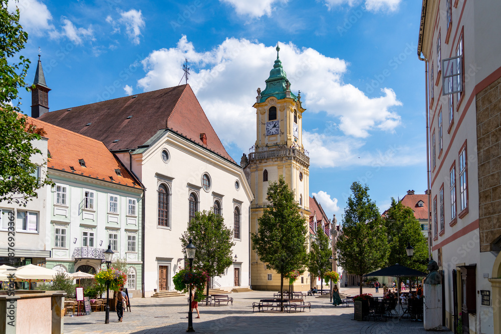 Obraz premium Visitors and locals alike enjoy a sunny day in Bratislava's historic Main Square (Hlavné námestie), which is anchored by the iconic yellow Old Town Hall tower and the white Jesuit Church.