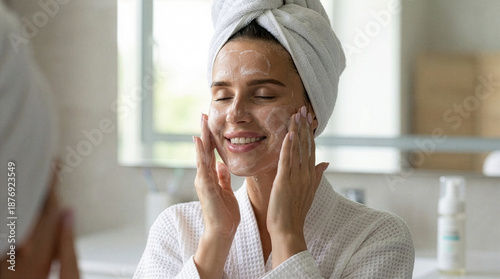 Woman applying skincare product in bathroom while looking in mirror and smiling with towel on head during morning routine