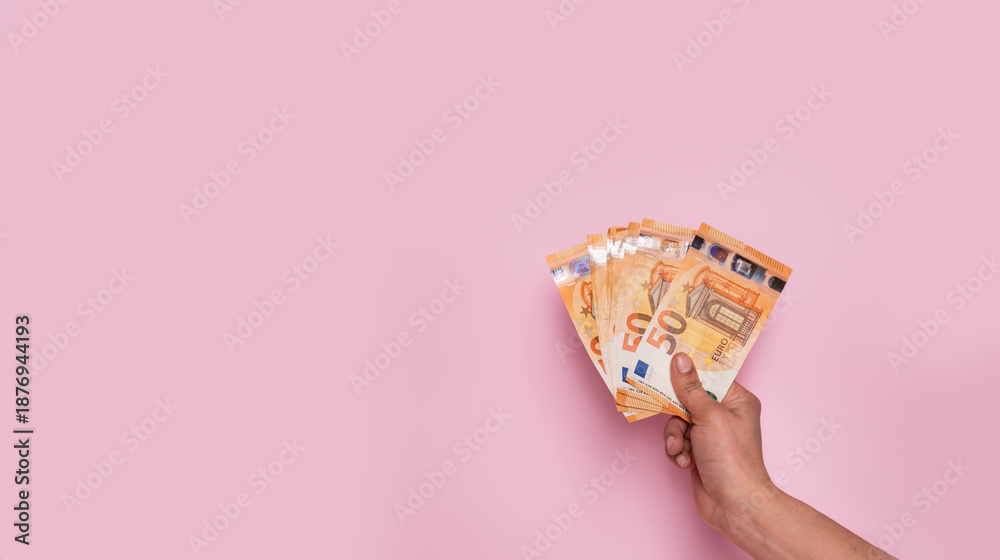 © Beatriz - Horizontal web banner of a hand holding euro banknotes on a pastel pink background with copy space. Earn money, invest and save, cash concept
