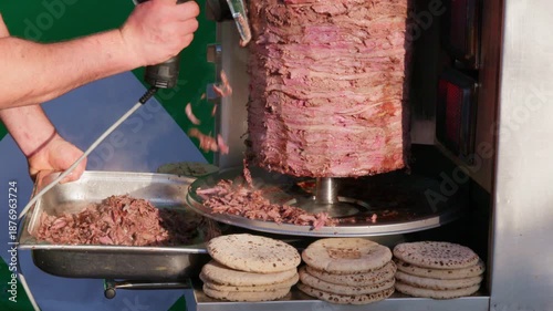 Street vendor slices hot shawarma doner from a vertical rotisserie with an electric knife as meat falls into a tray, fresh pita breads stacked in front, ready for fast service