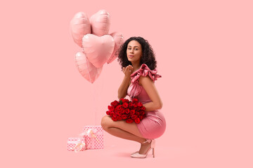 Beautiful young African-American woman with bouquet of roses and heart-shaped balloons blowing kiss on pink background. Valentine's Day celebration