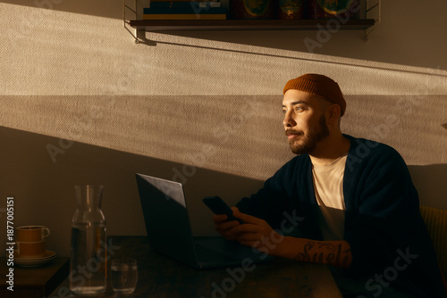 A focused man with a beard and beanie working on his laptop at a wooden table during golden hour. The warm sunset light creates dramatic shadows across the wall in a cozy indoor setting.