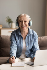 Diligent and inspired aged student. Home office portrait happy cheerful smart older lady in big headphones sit on sofa by laptop look at camera ready to take notes passionate about elearning education