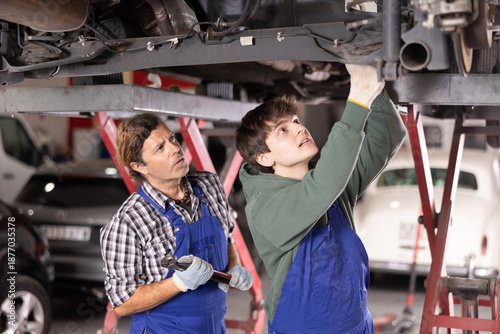 Foto Two focused professional mechanics, middle aged and young men, wearing blue overalls, repairing undercarriage of car raised on lift in auto repair shop