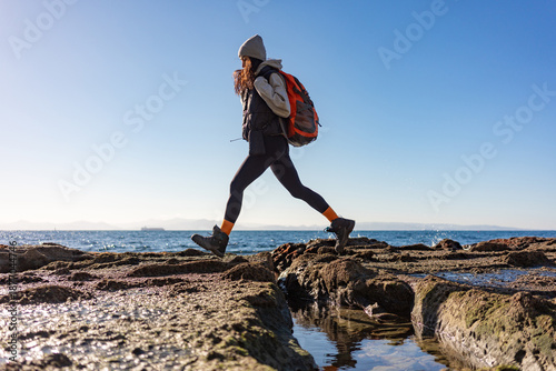 Foto Active female hiker with orange backpack jumping over rocky coastal terrain at l