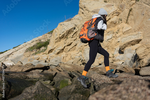 Φωτογραφία Female hiker with orange backpack carefully stepping across uneven coastal rocks
