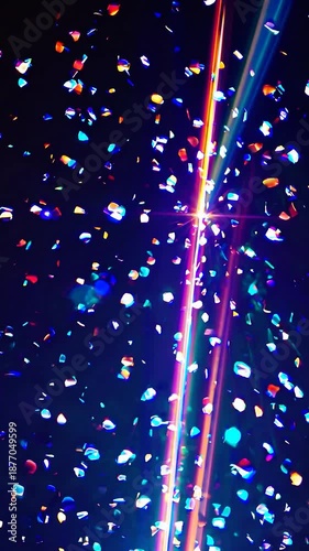 Shiny, iridescent confetti pieces floating down against a dark background. A bright, golden light source creates stunning rays and lens flare