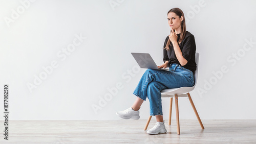 Tableau sur toile Troubled Caucasian woman sitting on chair with laptop computer, making mistake in online project, feeling stressed, thinking over solution to problem against white wall, copy space