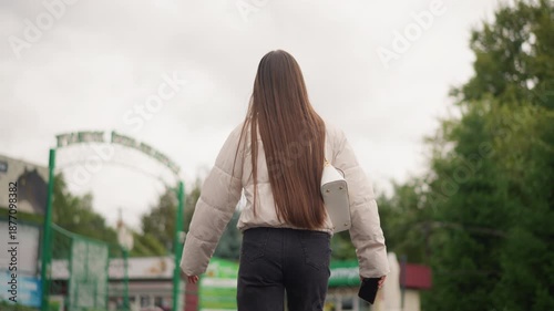 young woman walking toward park entrance under overcast sky, long braided hair in white jacket and jeans, walking away from camera past green gate and trees, shallow depth with soft bokeh, muted tones