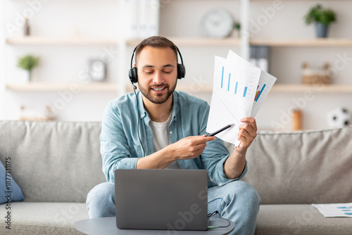 Photography Cheery young guy in headset making video call, showing documents at webcam, using laptop, working from home