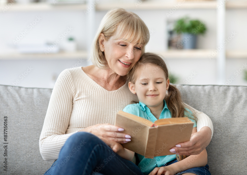 Fototapeta premium Family Pastimes. Lifestyle portrait of loving caring granny reading interesting book together with her cute little granddaughter, hugging and sitting on the couch at home in living room