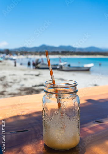 Refreshing drink in mason jar by the beach.  Cold beverage in a glass jar on a sunny beach with boats and blue sea in the background.