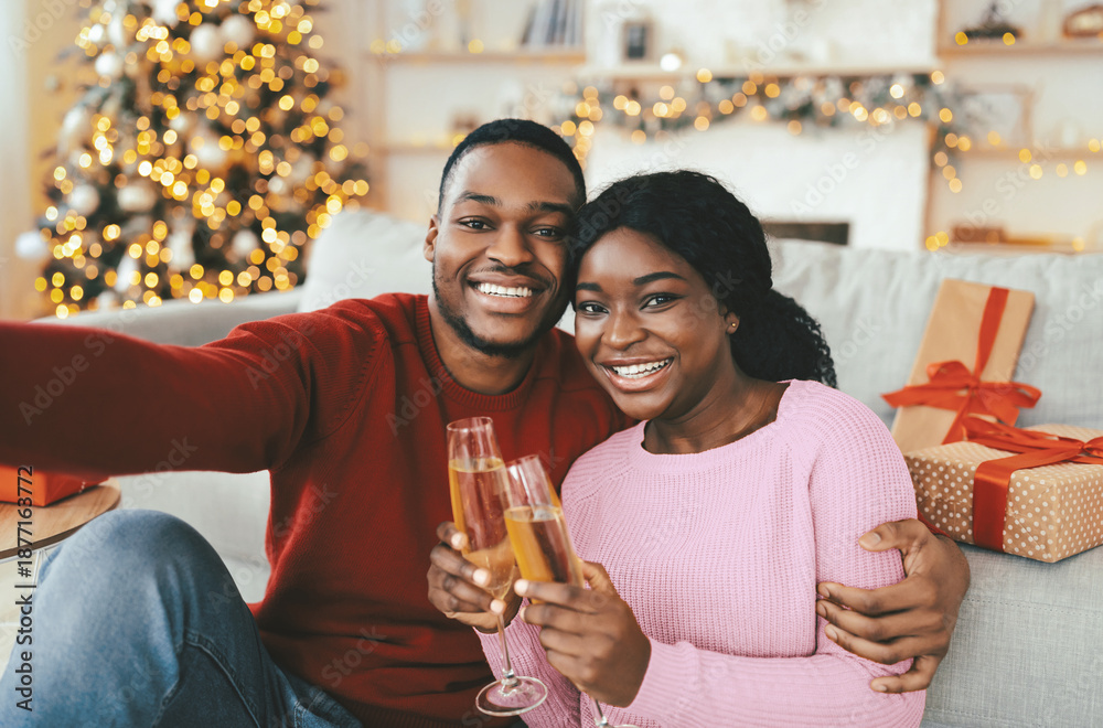 Obraz premium Two individuals smile and hold glasses of drink while sitting on a couch in a festive living room. There are holiday decorations and wrapped gifts nearby, creating a joyful scene.
