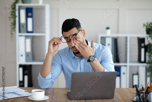Fotografía Young Arab businessman having tired irritated eyes, sitting at desk in front of laptop, exhausted from online work at modern office