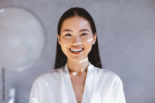 A woman smiles while wearing a white robe in a well-lit indoor room. Soft light filters through the space, highlighting her joyful expression and creating a warm atmosphere.