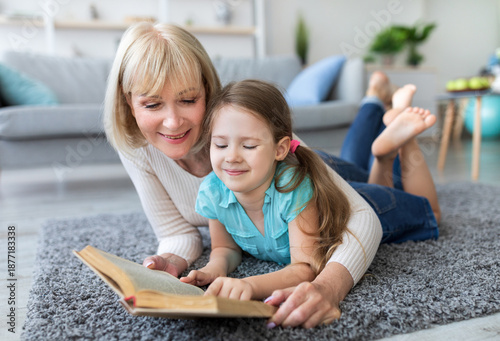 Bild auf Leinwand Loving grandmother teaching granddaughter holding book lying on rug floor carpet
