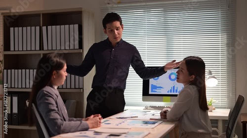 Three people are sitting at a desk with a computer monitor. A man is standing in front of the monitor