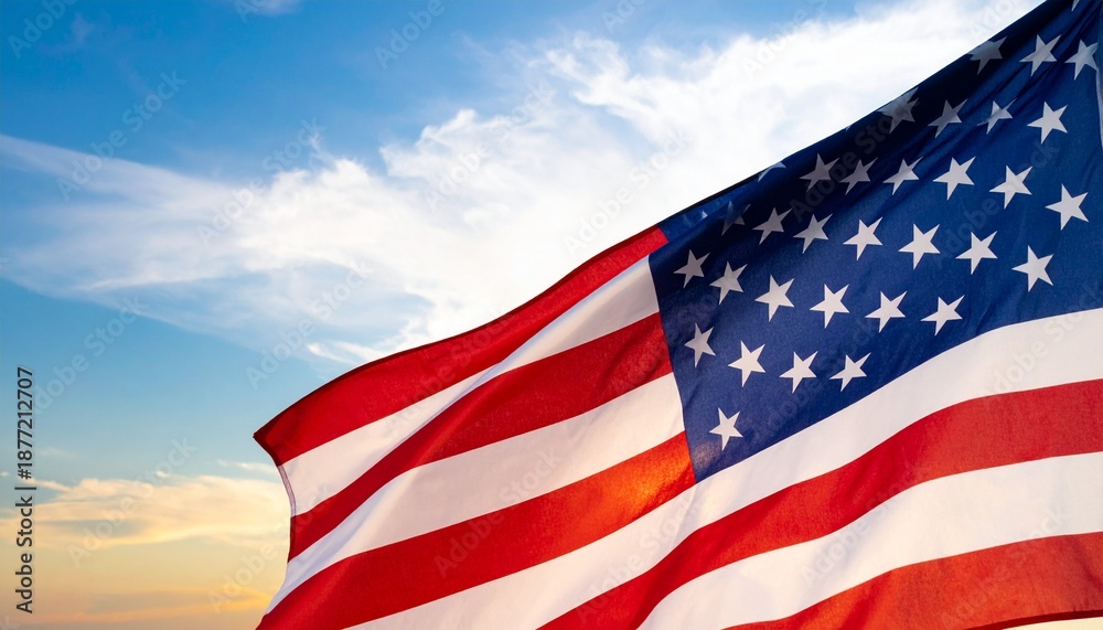 Fototapeta premium The American flag proudly waves against a vibrant blue sky with soft white clouds, illuminated by warm sunlight.