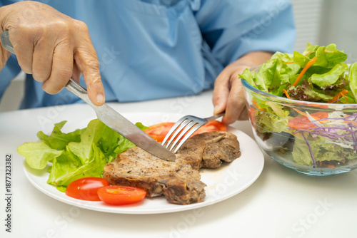 Asian elderly woman patient eating pork chop stake and vegetable salad for healthy food in hospital.