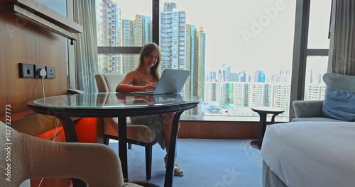 Young woman smiling and working on laptop, typing in a luxurious hotel room with a panoramic city skyline view. Remote work, digital nomad, business travel, modern lifestyle, urban productivity