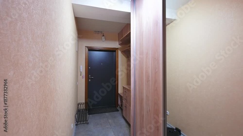 narrow hallway with wooden sliding doors, built-in shelving, dark door at the end, robotic vacuum cleaner on the floor, neutral-toned walls, recessed ceiling lights. Compact, functional space.