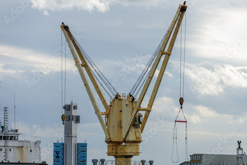 Photography Landscape view of shipboard deck crane preparing to lift heavy loads with cables and hanging hook dangling in midair against cloudy sky backdrop over a shipyard