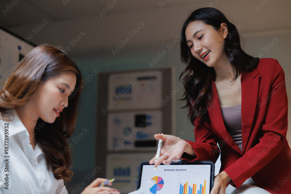 Fototapeta premium Two businesswomen collaborating on data analysis, reviewing Business Reports displayed on a digital tablet with colorful pie and bar charts in a professional office setting