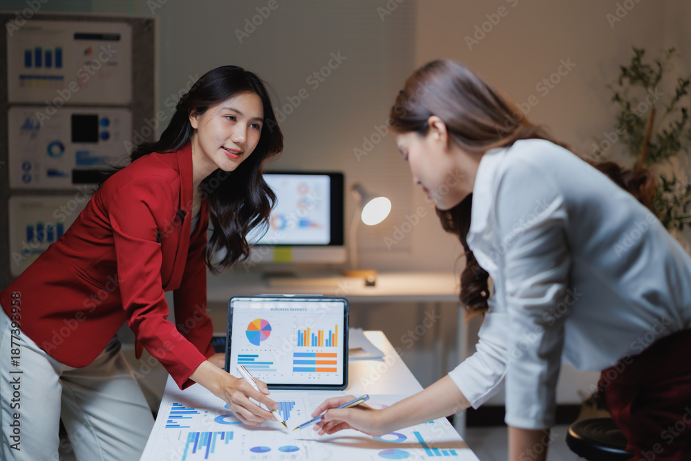 Fototapeta premium Two businesswomen discussing financial reports and pie charts during a late business meeting, analyzing market data and collaborating on a new project using a digital tablet in the office