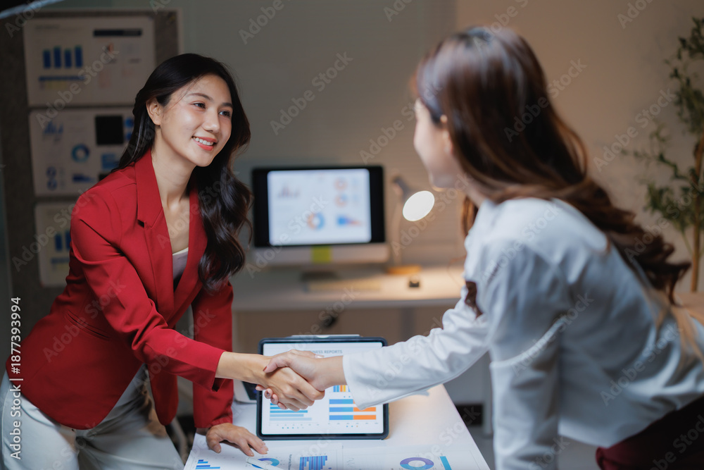 Fototapeta premium Two Asian businesswomen collaborating, sealing a deal with a handshake, showing teamwork and partnership in a modern office environment, with reports and tablet on the desk