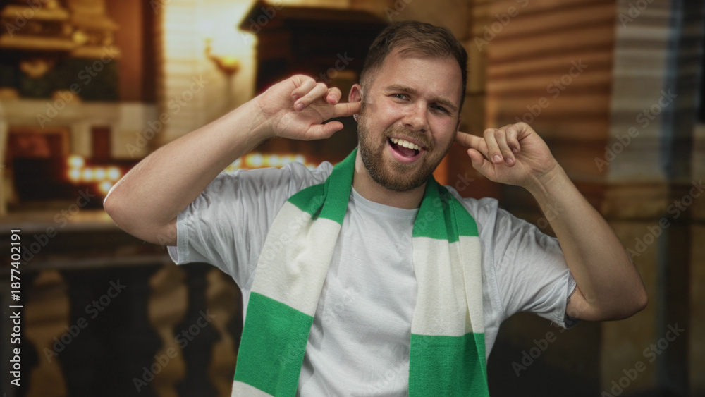 Obraz premium Man wearing green and white scarf plugs ears with fingers and smiles inside a church building, caucasian young fan in white tshirt; defiance.