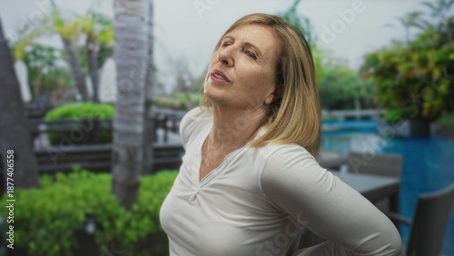 Φωτογραφία Middle age woman holds lower back and leans backward in green park by poolside chairs on sunny morning; discomfort