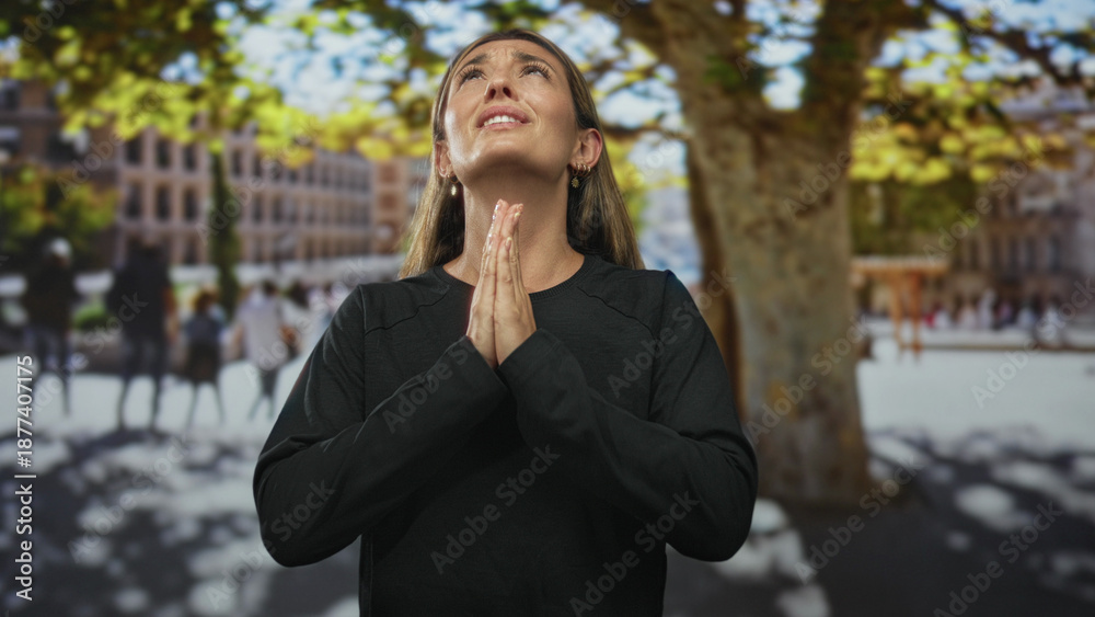 Fototapeta premium Young woman with hands pressed together praying on a sunlit street plaza beside a large plane tree and blurred buildings; hope gratitude faith.