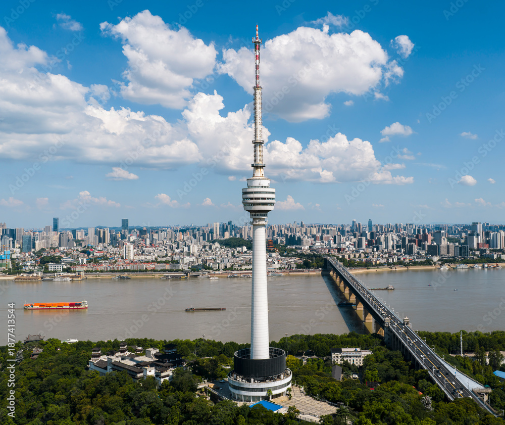 Obraz premium Wuhan TV Tower stands majestically by the Yangtze, surrounded by a bustling cityscape and serene river traffic, under a summer sky dotted with clouds. The tower, a beacon of technology, with sleek, fu