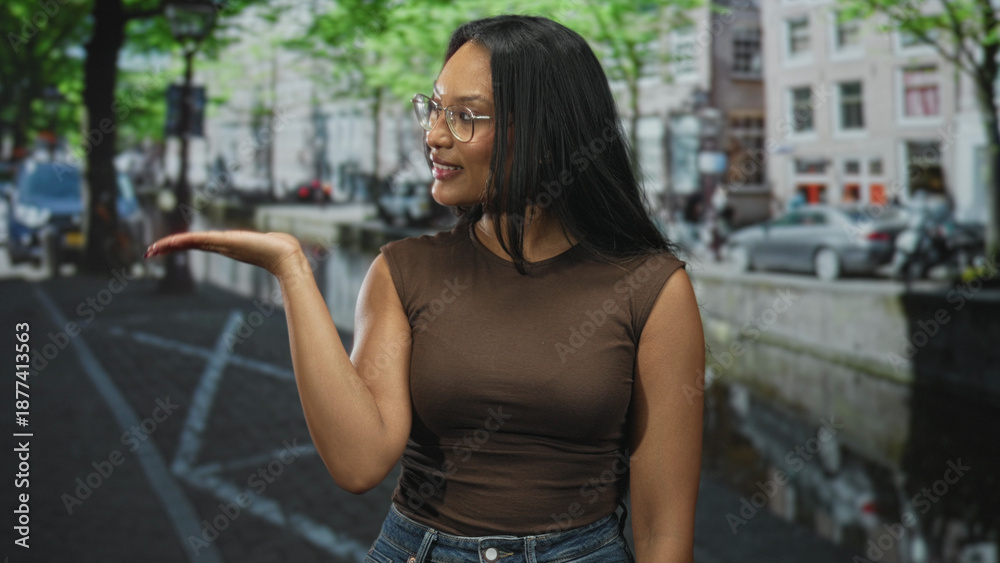 Fototapeta premium Woman points finger up on a canal street with glasses and brown top, presenting gesture by parked cars and canal water; confidence invitation.