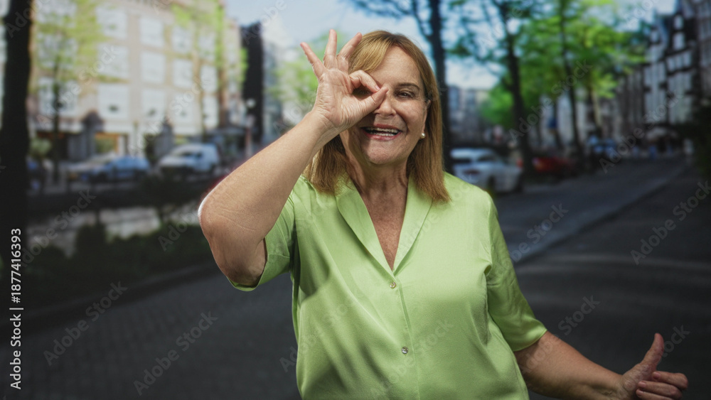 Fototapeta premium Older woman making ok sign with hand over eye on a city street by a canal, smiling and wearing a green blouse near parked cars; playful joy.