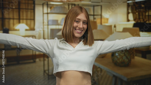 Young blonde woman smiling with arms outstretched in a stylish lamp shop, surrounded by various indoor light fixtures creating a warm and inviting atmosphere. Free Download Young blonde woman smiling with arms outstretched in a stylish lamp shop, surrounded by various indoor light fixtures creating a warm and inviting atmosphere. - GFXLoad
