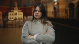 Woman standing in a church with arms crossed wearing a grey sweater looking serious, suggesting a religious context indoors with subdued lighting.