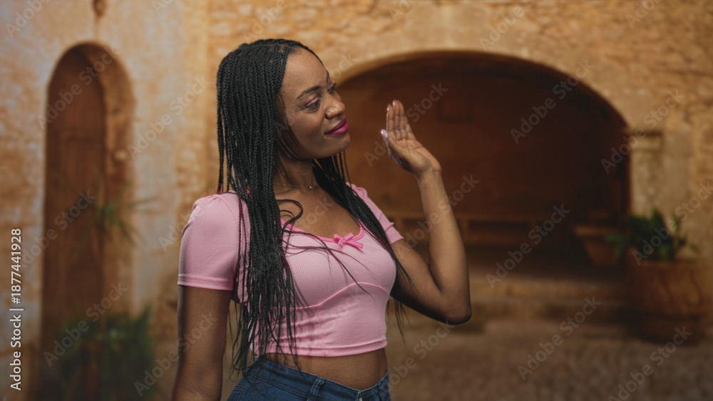 Fototapeta premium Black woman flips braided hair with raised hand, wearing pink shirt and visible midriff in a stone street courtyard; confidence attitude.