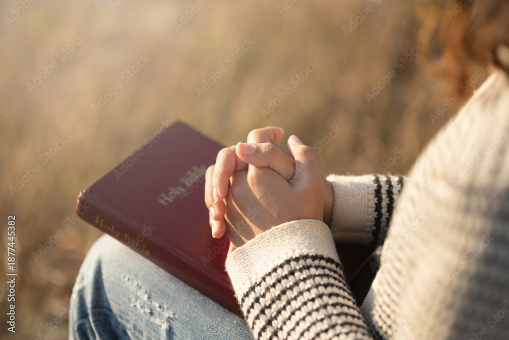Fototapeta premium A close-up image of a Christian woman clasping her hands in prayer over a red Bible, seeking love, hope, faith, and guidance through prayer and Bible reading outdoors during a tranquil sunset.
