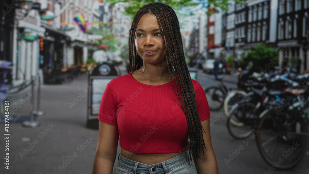 Naklejka premium Woman with braided hair and visible breasts standing on a city street in a red cropped shirt; quiet confidence.