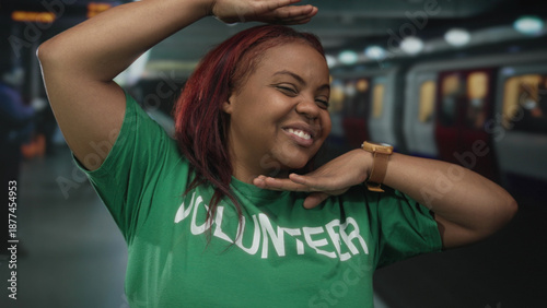 Volunteer in a green shirt smiles and frames her face with her hands beside a train in a building; playfulness community service volunteering.