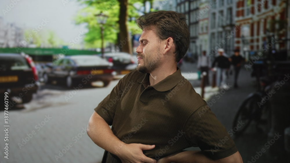 Fototapeta premium Man clutching right side and holding lower back on city street with backache near parked cars and bicycles; acute pain.